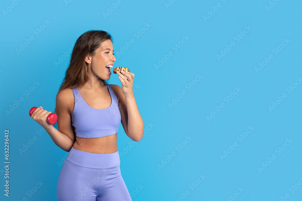 Glad millennial european lady in sportswear doing arm exercises with dumbbell and eating donut Glad millennial european lady in sportswear doing arm exercises with dumbbell and eating donut