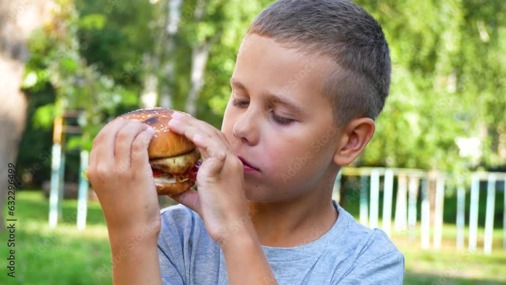 Cute 9-year-old boy eating a burger in the park in the summer. A hearty snack for a child, street food. The concept of healthy nutrition for children Cute 9-year-old boy eating a burger in the park in the summer. A hearty snack for a child, street food. The concept of healthy nutrition for children
