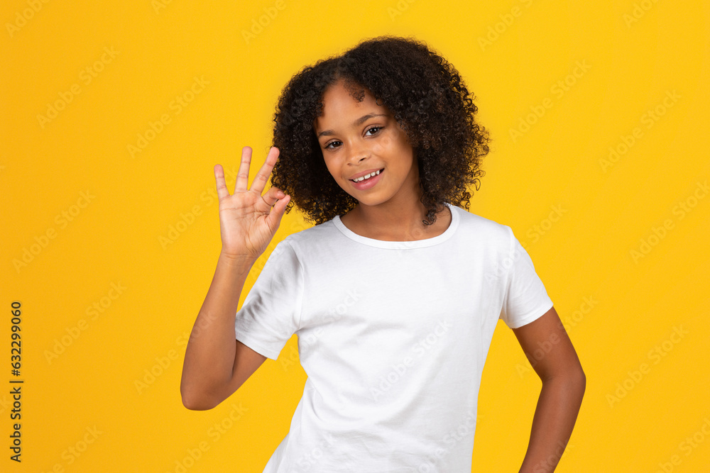 Happy teenage black pupil girl in white t-shirt shows ok hand sign, isolated on yellow background Happy teenage black pupil girl in white t-shirt shows ok hand sign, isolated on yellow background