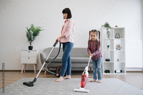 Brunette woman and tween girl utilizing different models of vacuum cleaners while removing dirt in living room. Diligent young homemakers sharing responsibilities of keeping apartment tidy. photo