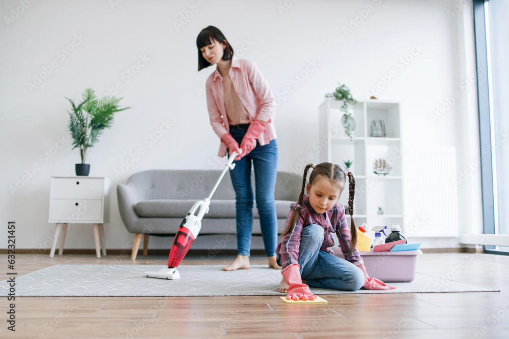 Helpful tween girl in rubber gloves polishing laminate floor while brunette woman vacuuming rug with electric appliance. Focused stay-at-home mom involving daughter in household chores at weekend.