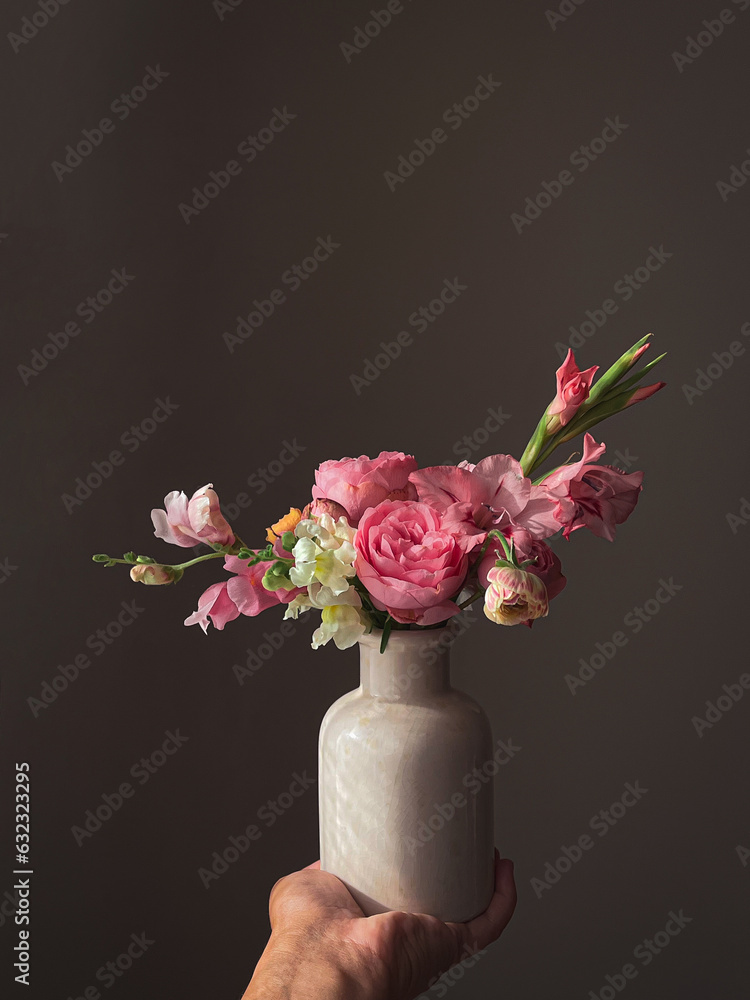 Hand holding beautiful pink flowers in vase in light on grey background. Stylish composition of snapdragon, roses, ranunculus,gladiolus. Flowers Still life. Floral vertical wallpaper