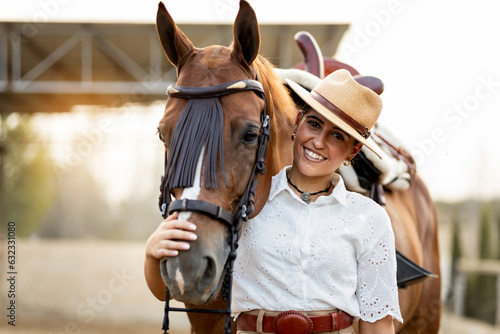 A cute girl in neat clothes poses caressing the muzzle of her brown horse while looking at the camera happily. The animal is of the Algo-Arabian breed. Young women riding horses.