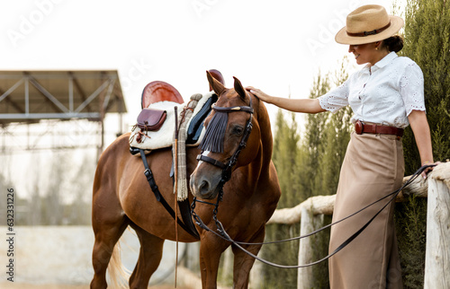 A cute girl in neat clothes is leaning against some fences resting while petting her brown horse outside the riding arena. The animal is of the algo-Arab breed. Young women riding horses.