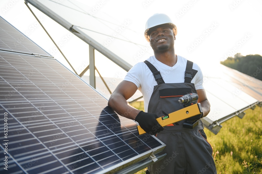 African american man in white helmet and grey overalls standing among rows of solar panels African american man in white helmet and grey overalls standing among rows of solar panels