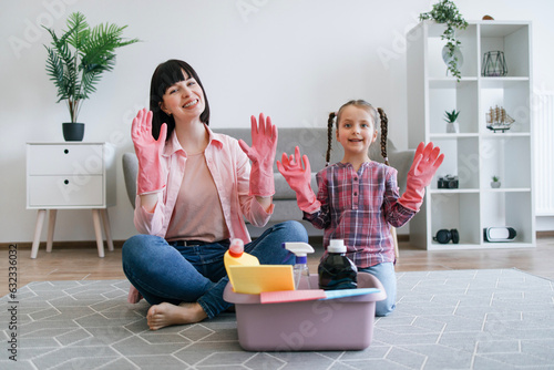 Portrait of smiling mother and daughter showing hands in rubber gloves while sitting near plastic tub with cleaning supplies. Cheerful homemakers getting ready for polishing and dusting off room. photo