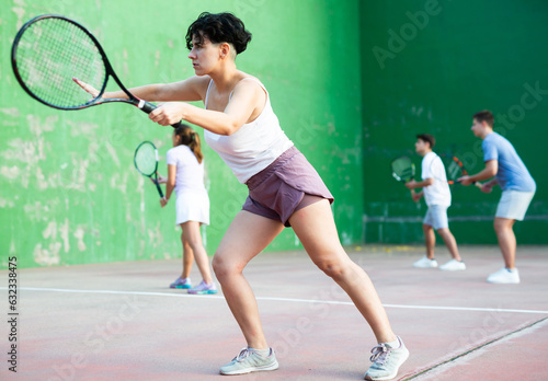 Sporty young Argentinian woman playing popular team match of frontenis at open-air fronton on summer day, ready to hit rubber ball with racquet © JackF