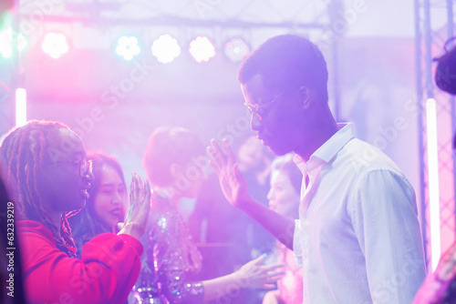African american couple showing hands moves while improvising dance battle in dark nightclub illuminated with lights. Young dancers partying at discotheque in crowded club