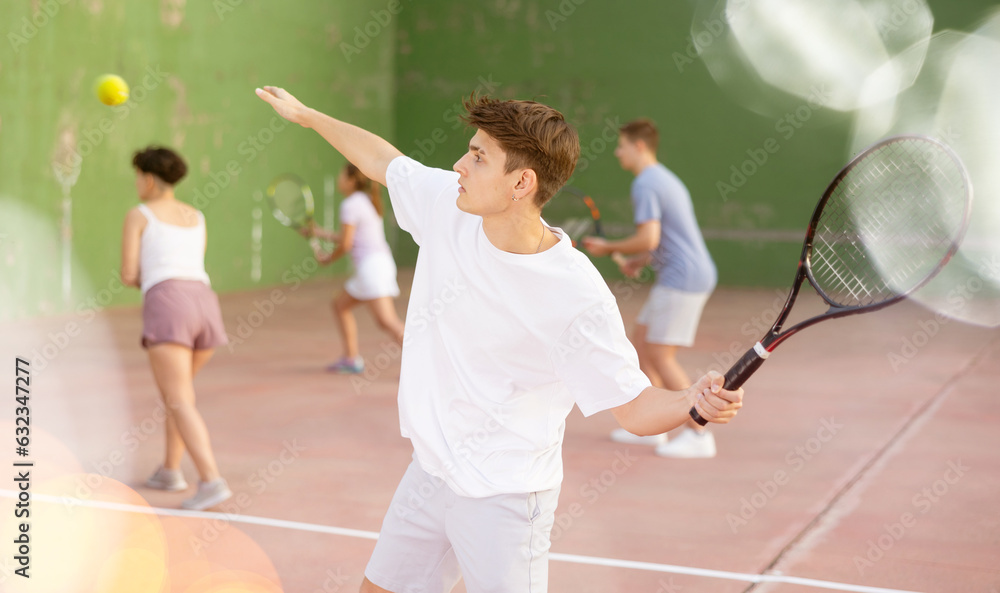 Concentrated young Latin American frontenis player swinging string racquet to hit ball on outdoor walled court on sunny summer day. Sport and active lifestyle concept