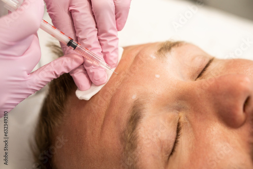 Men's cosmetology. Cosmetologist makes botulinum toxin beauty injection procedure in forehead wrinkles of handsome young man with beard in beauty salon.