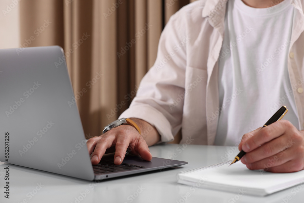 Man using laptop and writing something in notebook at table indoors, closeup Man using laptop and writing something in notebook at table indoors, closeup