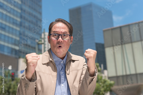 An elated middle aged man pumps both of his fists in success, looking at the camera. Reacting outside after being promoted to a senior managerial role. Outdoor city scene. photo