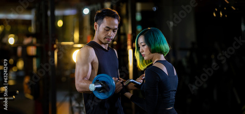 Asian woman is using easy bar as beginner in weight training on barbell for arm and core muscle inside gym with support from trainer to prevent injury dark background for exercising and workout