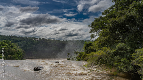 A stormy river winds between banks overgrown with lush tropical vegetation. Spray and mist over the water. Clouds in the blue sky. Iguazu National Park. Argentina.