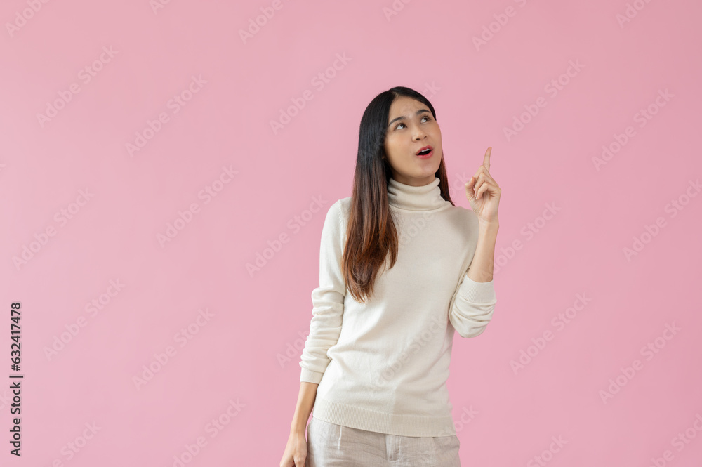 A cheerful Asian woman is looking upwards and pointing her finger up, isolated pink background. A cheerful Asian woman is looking upwards and pointing her finger up, isolated pink background.