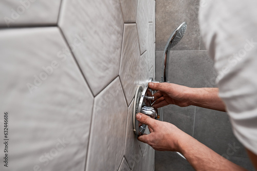Close up of man standing by the wall with ceramic tile and installing shower faucet with metal handle in apartment. Male plumber working on bathroom renovation at home. Plumbing works concept.