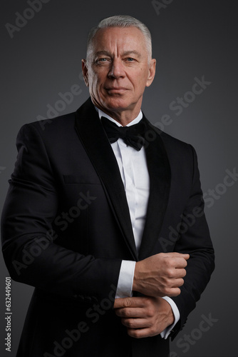 Сonfident and stylish senior man with grey hair wearing a black suit and bowtie on a grey background