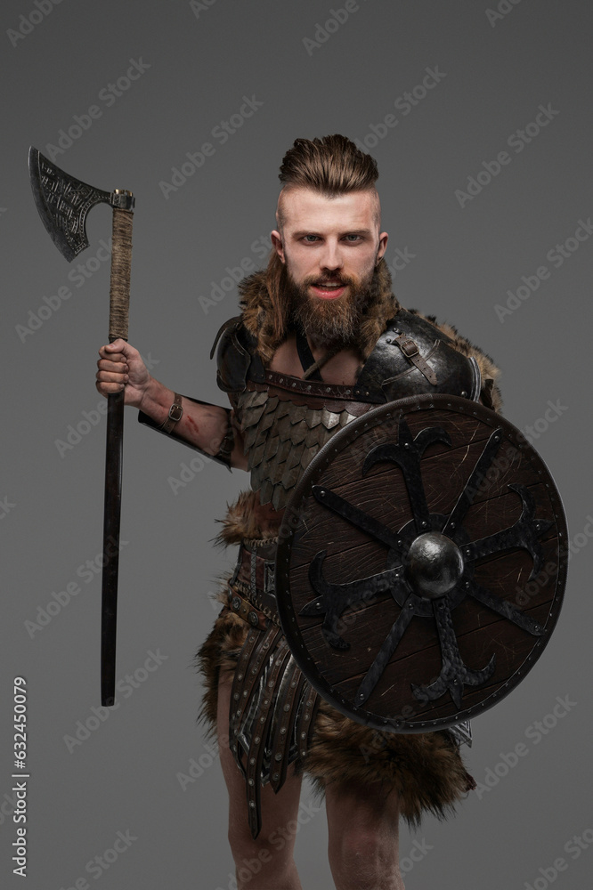 Naklejka premium Fierce bearded Viking dressed in fur and light armor wielding an axe against a gray background