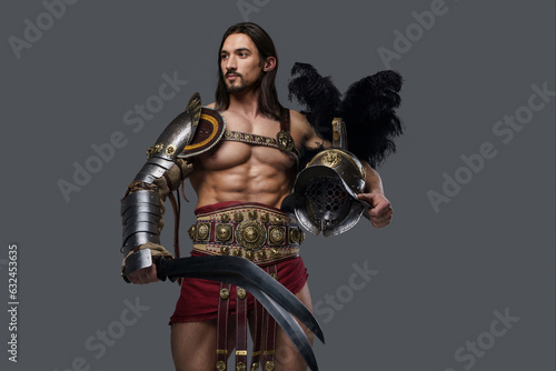 Powerful and attractive gladiator with a stylish beard and luscious locks wears ornate lightweight armor, clutching a gladius and feathered helmet as he stands confidently against a grey backdrop