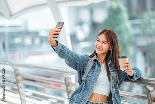 Fotografia Happy young asian woman taking selfie photo on mobile phone at city, female using smartphone camera for self portrait outside