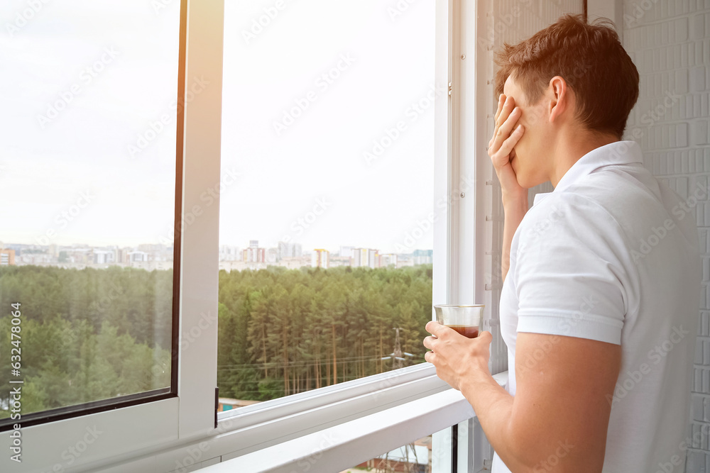 Man approaches open window overlooking forest on balcony drinking cup of coffee. City life of bachelor. Hot caffeinated drink for awakening, sunlight Man approaches open window overlooking forest on balcony drinking cup of coffee. City life of bachelor. Hot caffeinated drink for awakening, sunlight