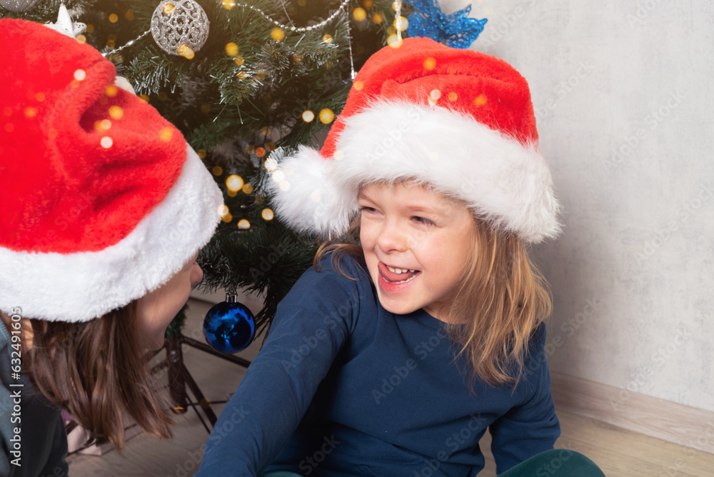 Happy cheerful children in santa hats under the Christmas tree. Christmas holiday. Real people, simple life. Happy cheerful children in santa hats under the Christmas tree. Christmas holiday. Real people, simple life.