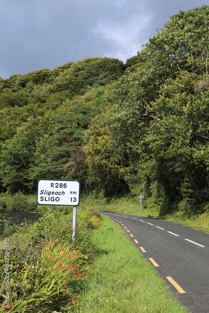 Rural road in County Leitrim, Ireland bordered by trees in foliage and Lough Gill Lake against backdrop of overcast sky with bilingual road sign visible in both English and Irish language Rural road in County Leitrim, Ireland bordered by trees in foliage and Lough Gill Lake against backdrop of overcast sky with bilingual road sign visible in both English and Irish language