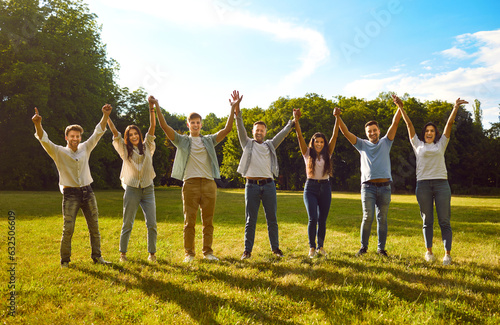 Group of friends having fun in summer park together. Team of happy cheerful young people standing in line on green grass lawn in the sun, holding hands up and smiling. Friendship, community concept