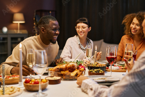 Laughing black man with flute of champagne bending over served table during chat with his friends while enjoying home party and dinner