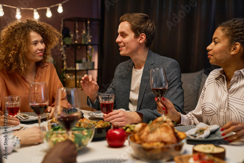 Young smiling man explaining something to his girlfriend by festive dinner while sitting among intercultural girls during communication