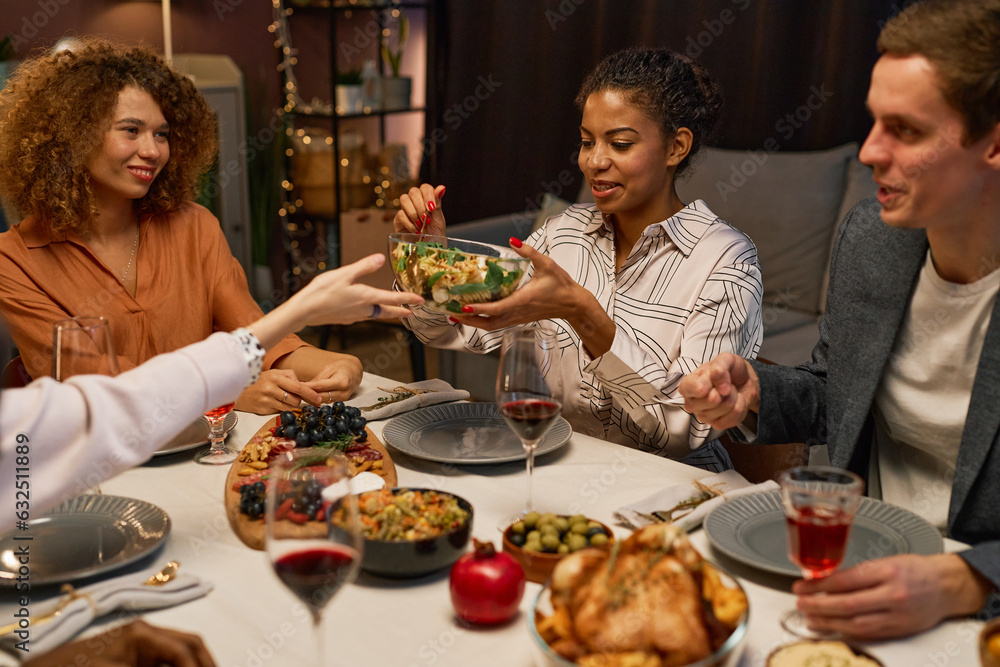 Young African American woman putting salad from glass bowl held by another girl over served festive table during home party