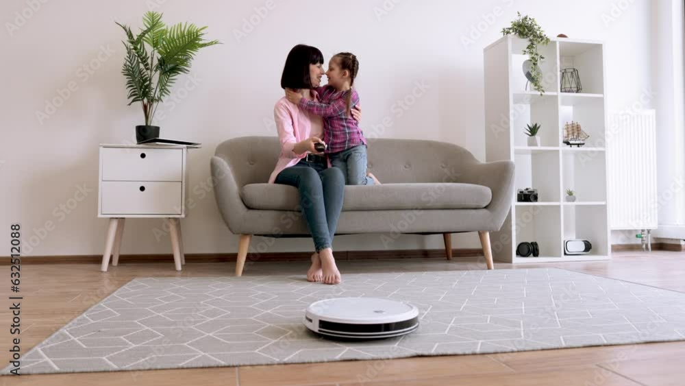 Full length view of smiling mother hugging sweet daughter on couch while turning on cleaning robot indoors. Cautious female parent involving kid into housekeeping routine with intelligent gadget. Full length view of smiling mother hugging sweet daughter on couch while turning on cleaning robot indoors. Cautious female parent involving kid into housekeeping routine with intelligent gadget.