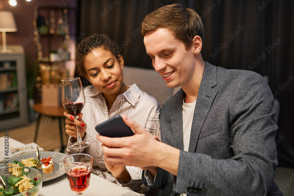 Young smiling man showing curious video in smartphone to his girlfriend with glass of red wine sitting next to him by served table during dinner