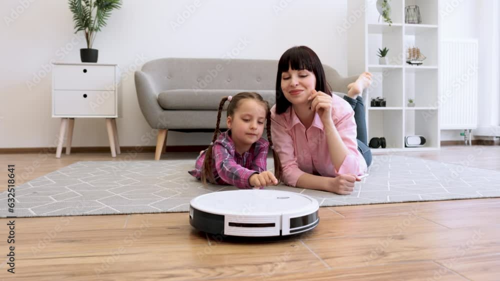 Happy mother and daughter pressing button on white robotic vacuum while lying on carpet in living room. Cheerful young ladies doing collaborative exercises on house during sunny weekend daytime. Happy mother and daughter pressing button on white robotic vacuum while lying on carpet in living room. Cheerful young ladies doing collaborative exercises on house during sunny weekend daytime.