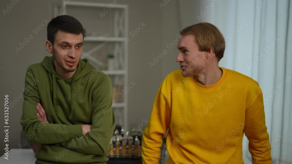 Two young relaxed men talking leaning on table smiling. Zoom in portrait of confident Caucasian male friends chatting enjoying leisure on weekend in living room Two young relaxed men talking leaning on table smiling. Zoom in portrait of confident Caucasian male friends chatting enjoying leisure on weekend in living room