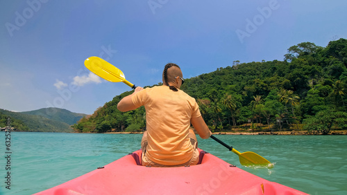 Young man with sunglasses and hat rows pink plastic canoe along sea against green hilly islands with wild jungles. Traveling to tropical countries. Strong guy is sailing on kayak in ocean, back view. © ivandanru