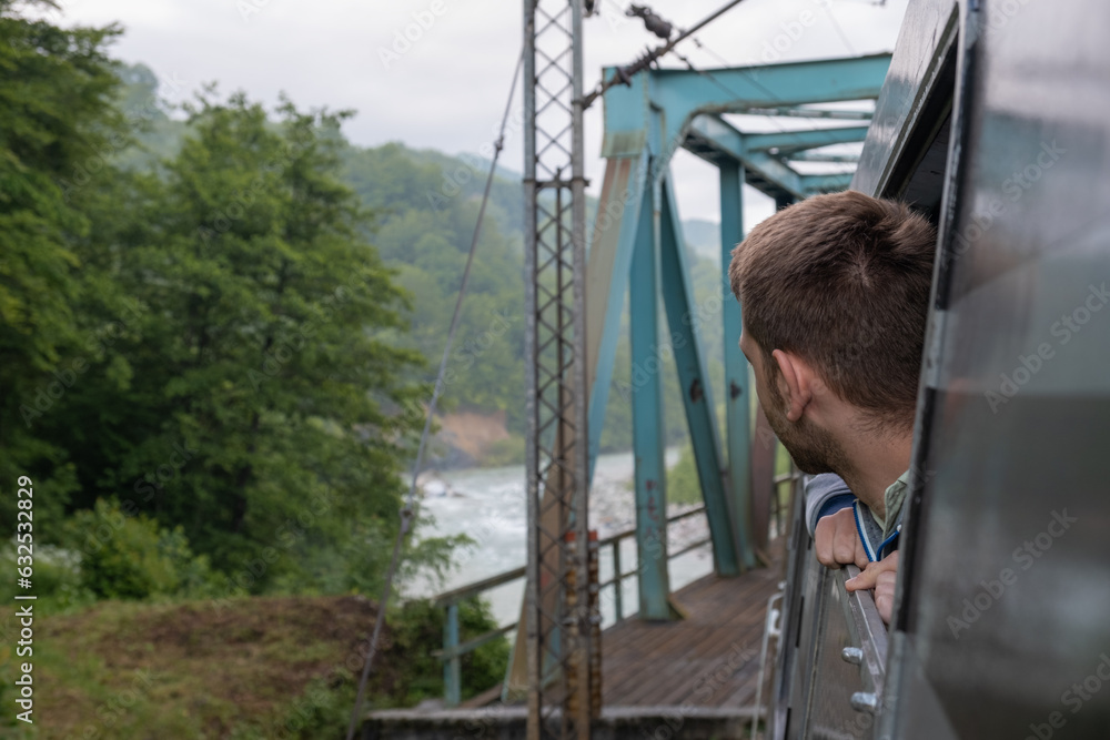 Young man looking out the train window while train cross the river over bridge. Male traveling by railway through Balkans