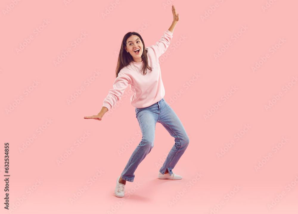 Happy young brunette, dressed in warm soft pink jumper and blue jeans, is fooling around with her arms outstretched and trying to keep her balance. Life-size studio photograph on isolated background. Happy young brunette, dressed in warm soft pink jumper and blue jeans, is fooling around with her arms outstretched and trying to keep her balance. Life-size studio photograph on isolated background.