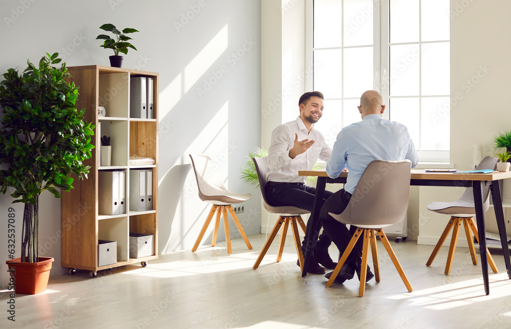 Two business men sitting at the desk of workplace talking and discussing company finance in office. Male company employers considering new projects, analyzing company. Team work concept. Two business men sitting at the desk of workplace talking and discussing company finance in office. Male company employers considering new projects, analyzing company. Team work concept.