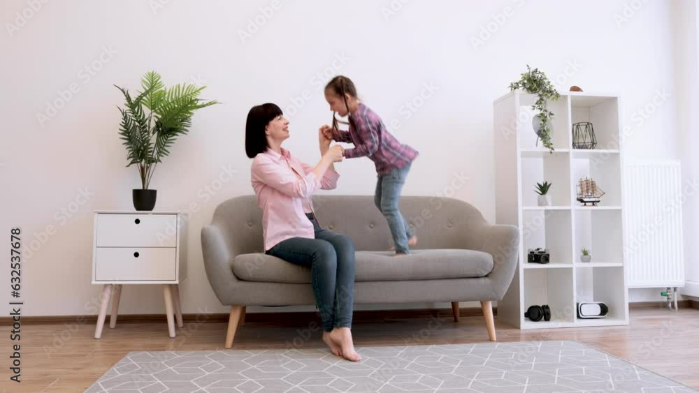 Charming woman holding child's hands while joyful girl jumping barefoot on soft sofa in spacious living room. Excited female parent turning mother-daughter time into engaging physical exercise. Charming woman holding child's hands while joyful girl jumping barefoot on soft sofa in spacious living room. Excited female parent turning mother-daughter time into engaging physical exercise.