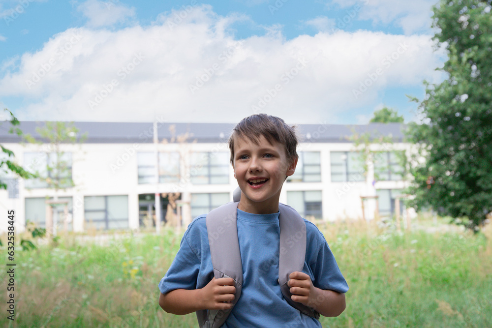 an 8-year-old boy stands in front of the school. a child in a blue T-shirt and a gray backpack. graduation or back to school. the boy goes to school through the park. an 8-year-old boy stands in front of the school. a child in a blue T-shirt and a gray backpack. graduation or back to school. the boy goes to school through the park.