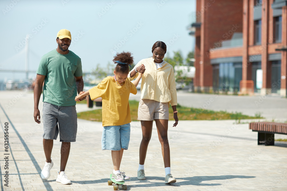 Parents holding hands with their child and teaching her to skateboarding in the park Parents holding hands with their child and teaching her to skateboarding in the park