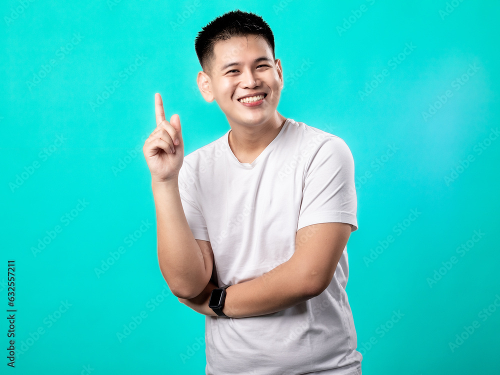 A portrait of an Asian man wearing a white t-shirt, posing as if having a brilliant idea with his finger pointing upwards, isolated with a turquoise background. A portrait of an Asian man wearing a white t-shirt, posing as if having a brilliant idea with his finger pointing upwards, isolated with a turquoise background.