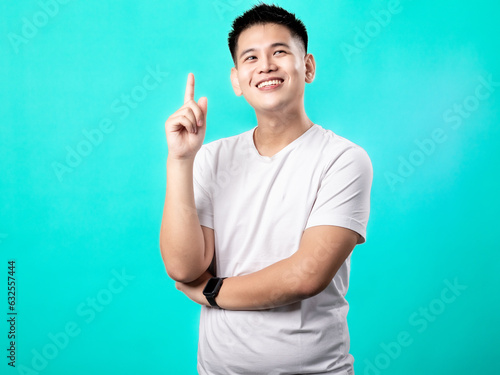 A portrait of an Asian man wearing a white t-shirt, posing as if having a brilliant idea with his finger pointing upwards, isolated with a turquoise background.
