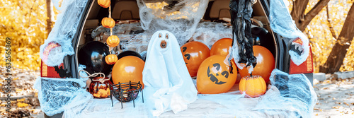 cute apricot poodle dog in ghost costume sits in trunk of car decorated for Halloween with cobwebs, orange balloons, pumpkins and sweets, the outdoor creative activity concept in autumn in October