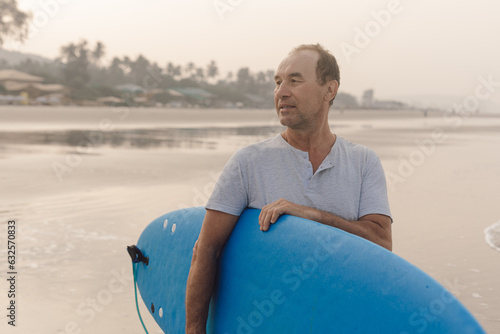 Canvas Print Confident mature surfer walking along empty shore at sunset, carrying blue surfboard under armpit and looking aside