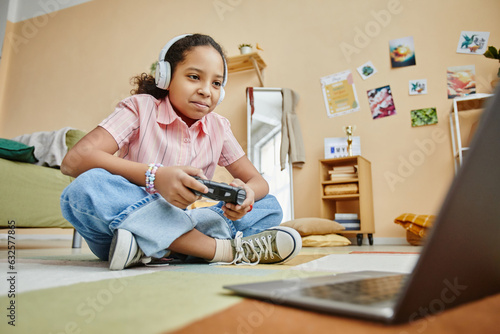 Happy schoolgirl with joystick pressing buttons and looking at laptop screen while sitting on the floor and playing video game at home