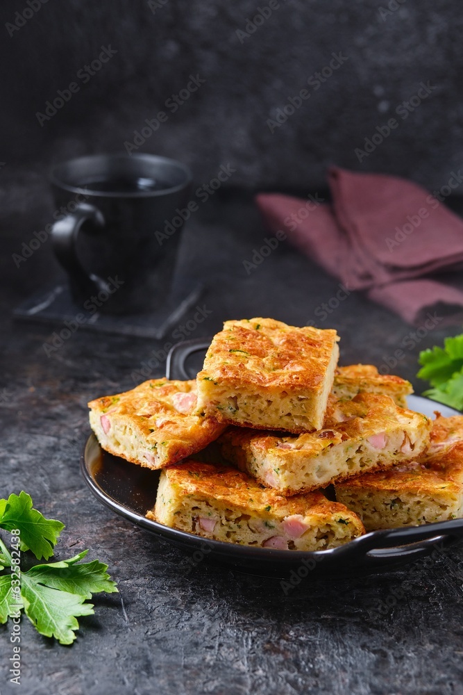 Sliced snack cake with zucchini, ham and cheese on a black plate on a dark concrete background. Recipes for zucchini. Sliced snack cake with zucchini, ham and cheese on a black plate on a dark concrete background. Recipes for zucchini.