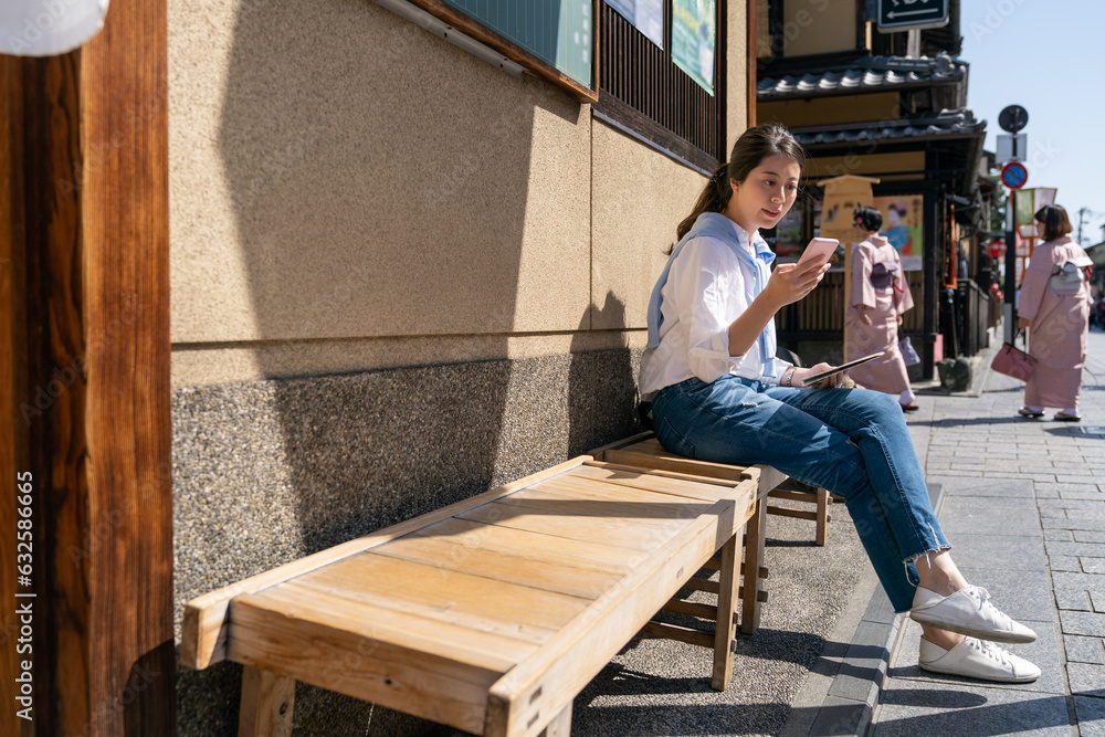 asian chinese female traveler consulting online travel info on phone while taking break on a bench on Gion Hanamikoji Street in Kyoto japan with two girls wearing kimono at background