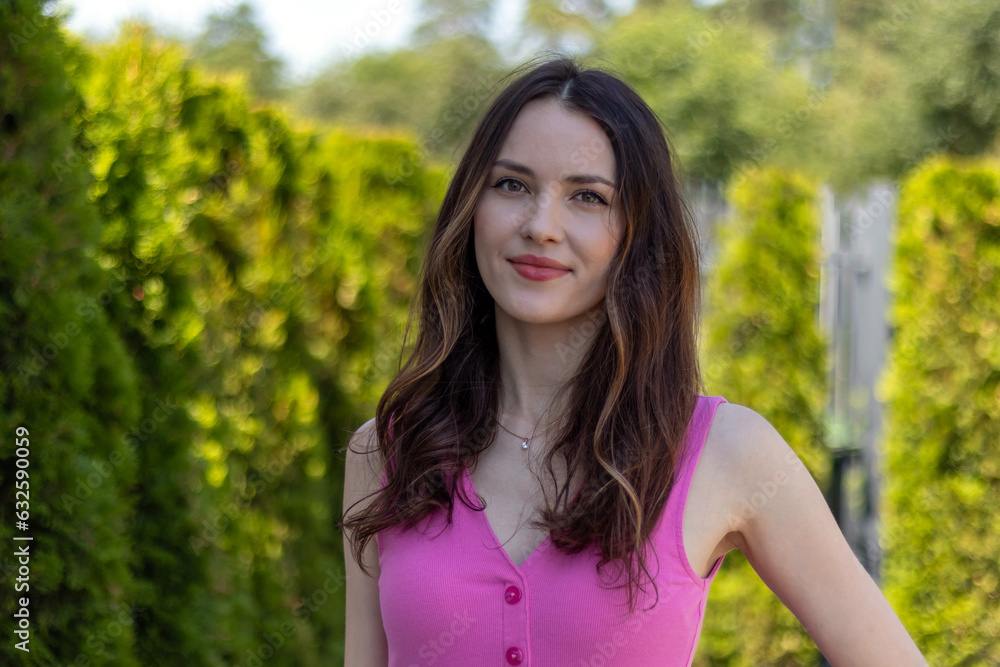 Portrait of a beautiful young woman model with wavy brunette hair in a pink dress against a background of green bushes Portrait of a beautiful young woman model with wavy brunette hair in a pink dress against a background of green bushes